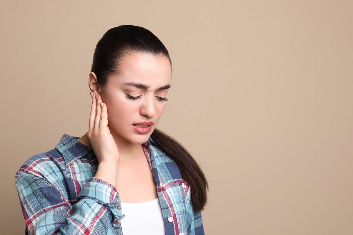 Young woman suffering from ear pain on beige background.